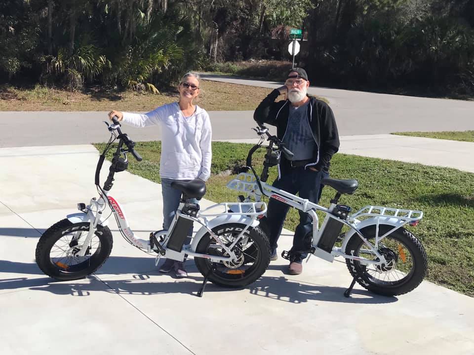 Two smiling customers posing with a DJ Folding Step Thru fat tire e-bike and a DJ Folding Bike fat tire e-bike on a driveway