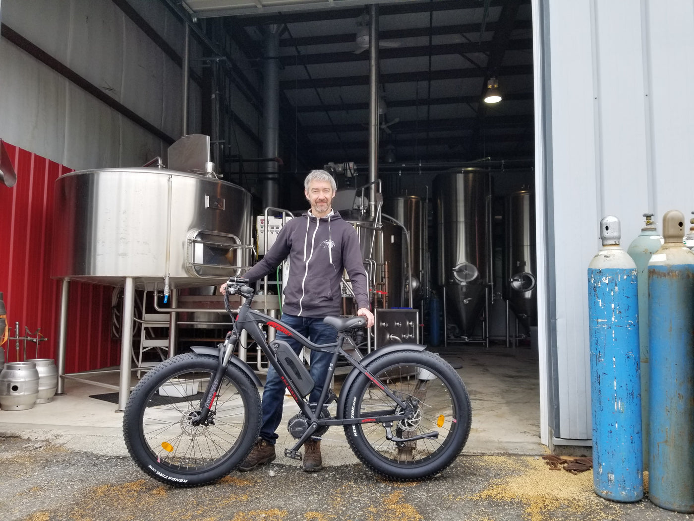 A smiling man poses with a DJ Fat Bike Mid-Drive fat tire e-bike in front of brewing equipment