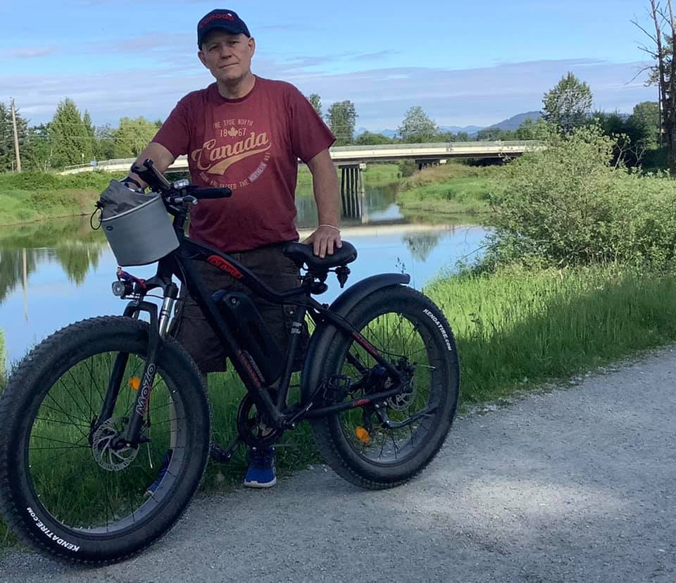 A smiling man poses with a DJ Fat Bike fit tire e-bike on a paved path in front of a river