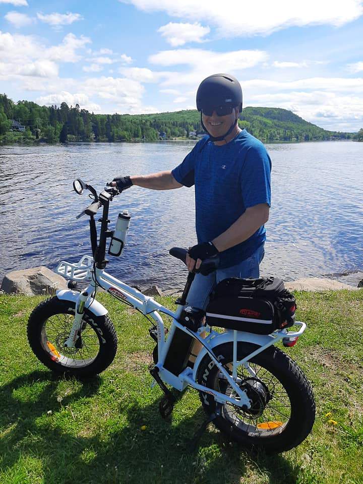 Smiling customer posing in front of a lake with a DJ Folding Bike fat tire e-bike equipped with trunk bag and water bottle holder